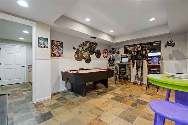 playroom featuring a tray ceiling, stone finish floor, recessed lighting, and a wainscoted wall