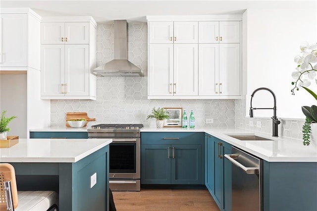 kitchen featuring wall chimney range hood, appliances with stainless steel finishes, a sink, blue cabinetry, and light countertops