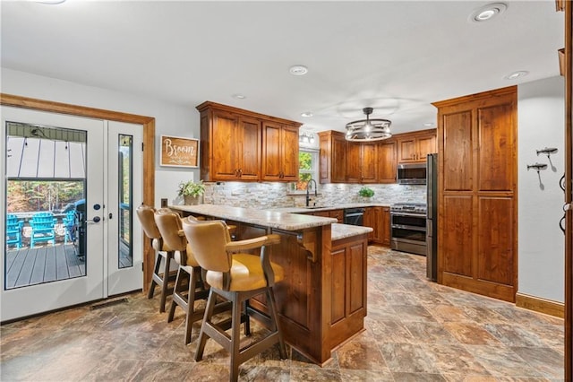 kitchen featuring stainless steel appliances, a peninsula, a sink, brown cabinets, and tasteful backsplash