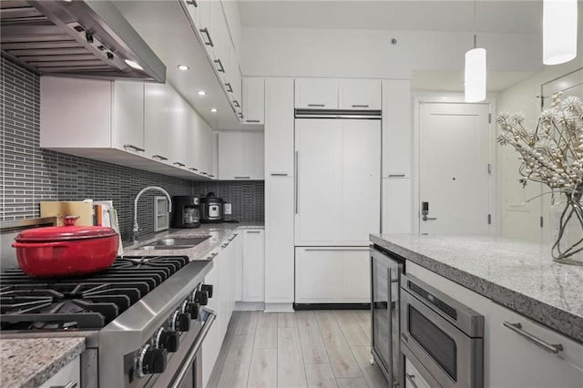 kitchen with a sink, wall chimney range hood, built in appliances, white cabinets, and backsplash