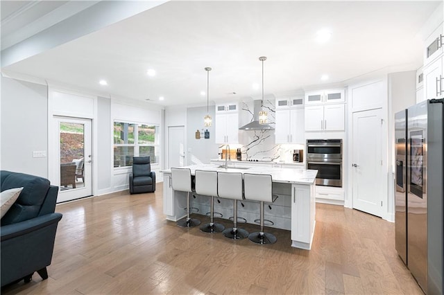 kitchen featuring wall chimney exhaust hood, hanging light fixtures, a kitchen island with sink, white cabinets, and appliances with stainless steel finishes