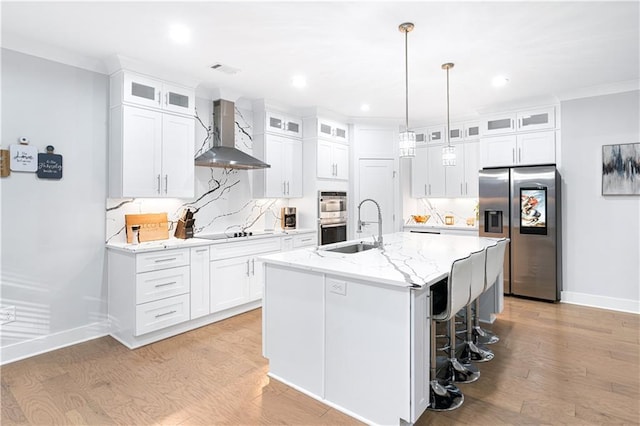 kitchen featuring appliances with stainless steel finishes, white cabinetry, wall chimney range hood, and a kitchen island with sink