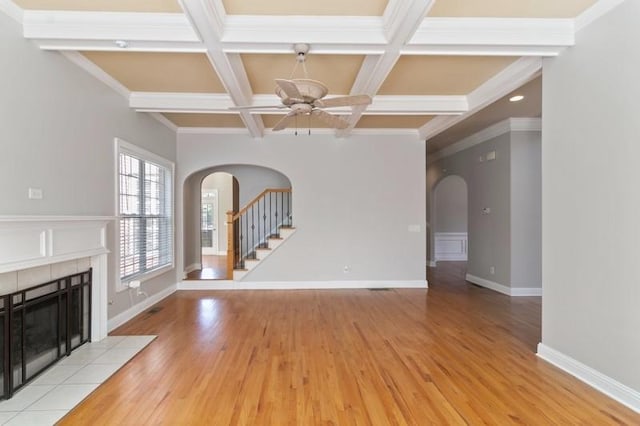 unfurnished living room with beam ceiling, a tiled fireplace, ceiling fan, light wood-type flooring, and crown molding