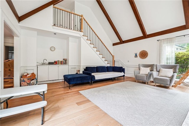 living room featuring high vaulted ceiling, stairway, beamed ceiling, wainscoting, and light wood finished floors