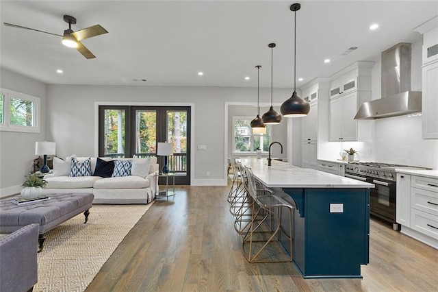 kitchen featuring wall chimney range hood, a breakfast bar, a kitchen island with sink, white cabinetry, and range with two ovens