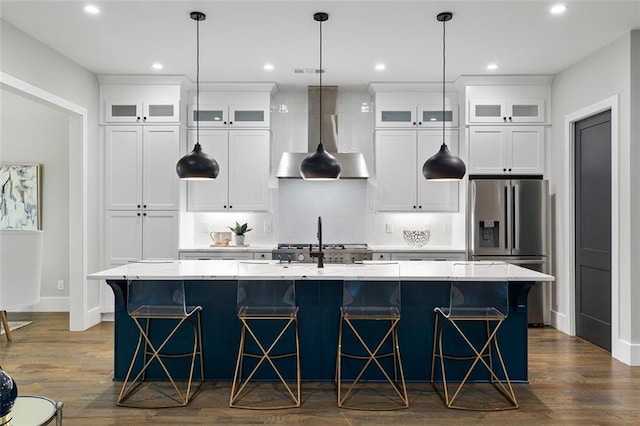 kitchen featuring wall chimney range hood, dark wood-type flooring, stainless steel fridge, a kitchen island with sink, and white cabinets