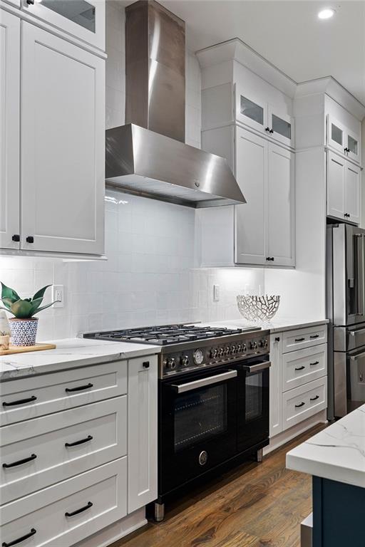 kitchen featuring range with two ovens, stainless steel refrigerator with ice dispenser, wall chimney range hood, and white cabinets