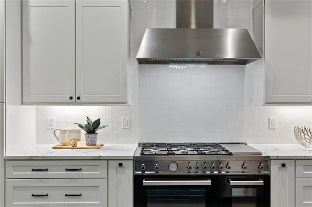 kitchen with wall chimney range hood, range, light stone counters, white cabinets, and decorative backsplash