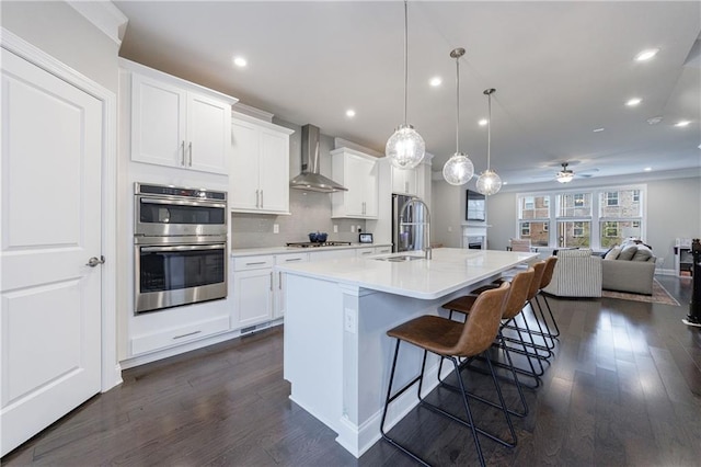 kitchen featuring appliances with stainless steel finishes, an island with sink, white cabinets, hanging light fixtures, and wall chimney range hood