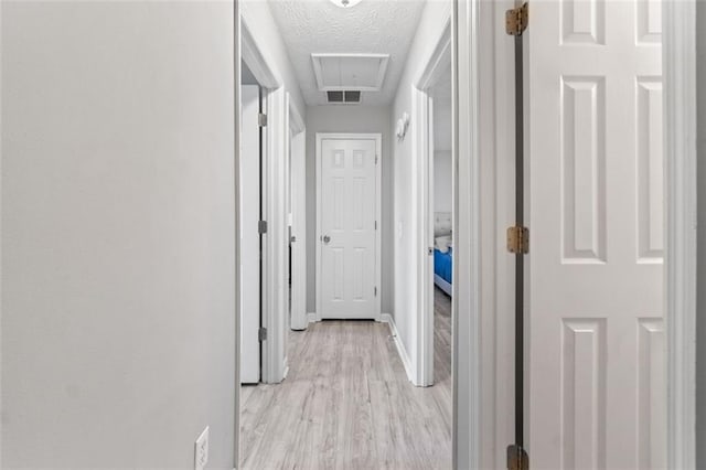 corridor featuring visible vents, a textured ceiling, light wood-style floors, baseboards, and attic access