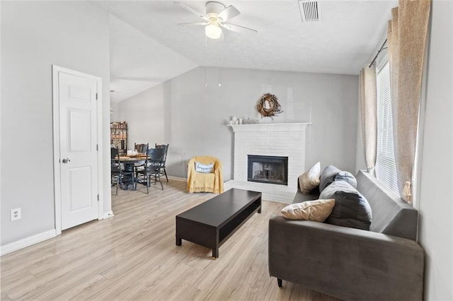 living area with visible vents, light wood-style flooring, a ceiling fan, a brick fireplace, and vaulted ceiling