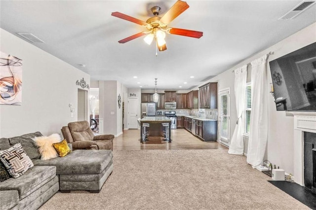 living area featuring a fireplace with flush hearth, a ceiling fan, visible vents, and light carpet