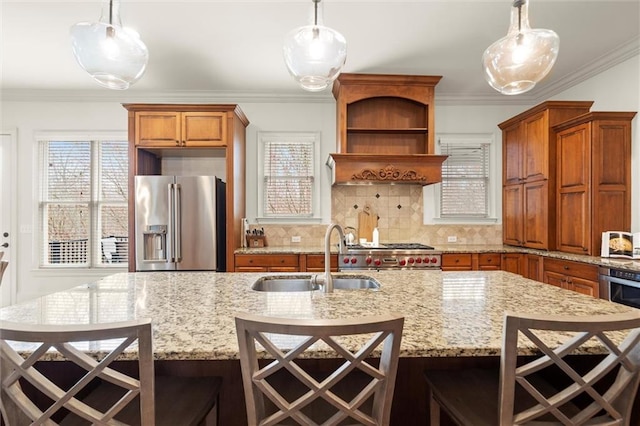 kitchen with high end fridge, ornamental molding, gas range, a sink, and an island with sink