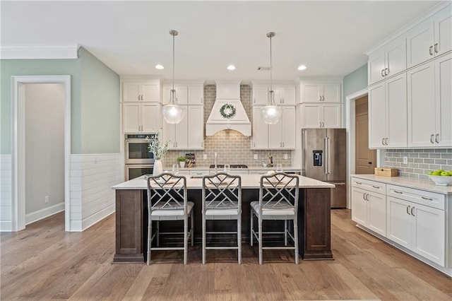 kitchen with a kitchen island with sink, stainless steel appliances, custom range hood, white cabinets, and decorative light fixtures