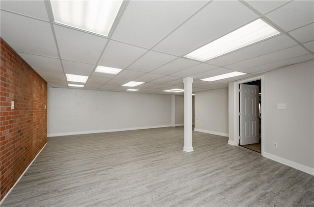 finished below grade area with a paneled ceiling, brick wall, baseboards, and wood finished floors