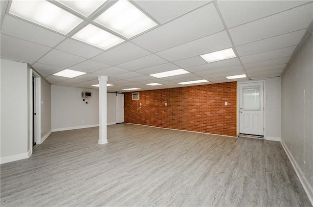 finished below grade area with a paneled ceiling, brick wall, light wood-style flooring, and baseboards