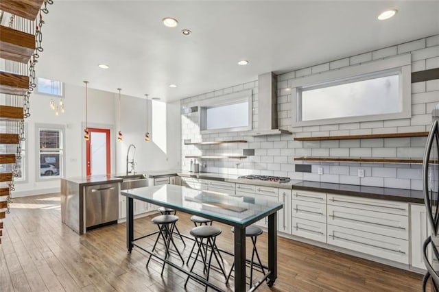 kitchen with a peninsula, stainless steel appliances, wall chimney range hood, backsplash, and open shelves