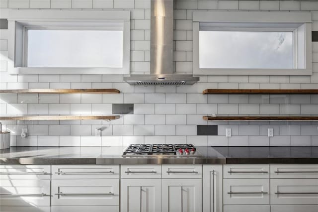 kitchen with stainless steel gas cooktop, stainless steel countertops, backsplash, white cabinets, and wall chimney exhaust hood