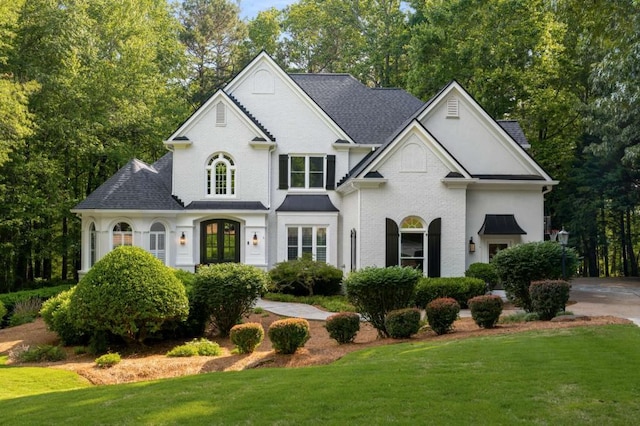 view of front facade with a front yard, brick siding, roof with shingles, and view of scattered trees