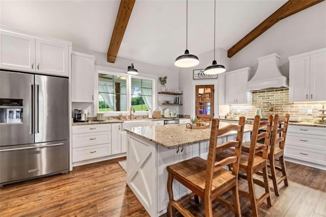 kitchen with a kitchen island, stainless steel refrigerator with ice dispenser, lofted ceiling with beams, white cabinets, and premium range hood