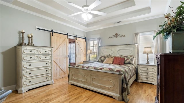 bedroom with light wood-type flooring, a tray ceiling, ceiling fan, crown molding, and a barn door