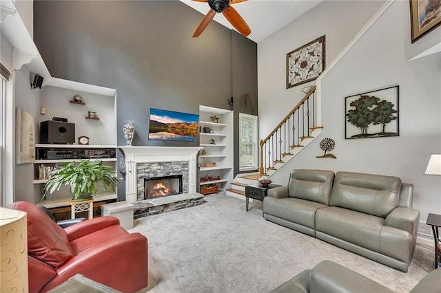 carpeted living room with stairs, a towering ceiling, a stone fireplace, built in shelves, and a ceiling fan