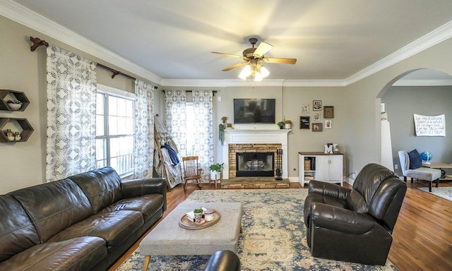 living room featuring arched walkways, ceiling fan, a stone fireplace, wood finished floors, and crown molding