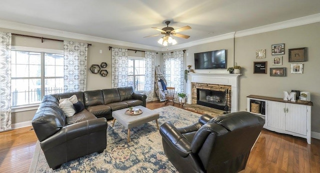 living area featuring baseboards, dark wood finished floors, a ceiling fan, ornamental molding, and a stone fireplace