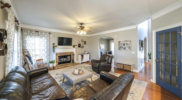 living area with arched walkways, a stone fireplace, wood finished floors, and crown molding