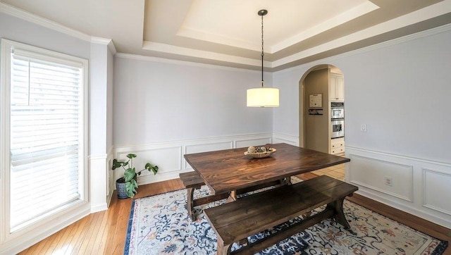 dining room featuring arched walkways, a raised ceiling, wainscoting, ornamental molding, and wood finished floors