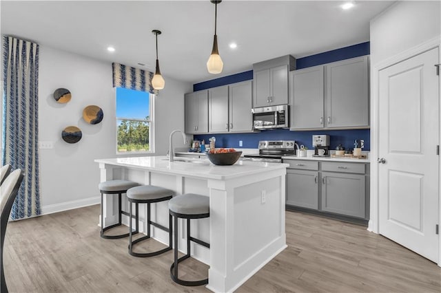 kitchen featuring appliances with stainless steel finishes, gray cabinetry, a sink, a center island with sink, and light wood-style flooring