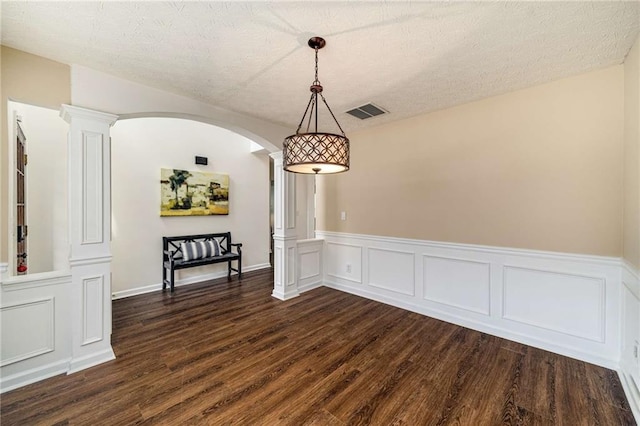 unfurnished dining area featuring decorative columns, visible vents, arched walkways, wainscoting, and dark wood finished floors