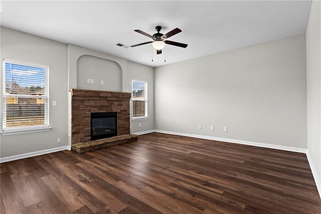 unfurnished living room with ceiling fan, visible vents, a stone fireplace, baseboards, and dark wood-style floors