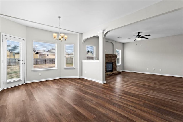 unfurnished living room featuring a stone fireplace, ceiling fan with notable chandelier, baseboards, and dark wood-style floors