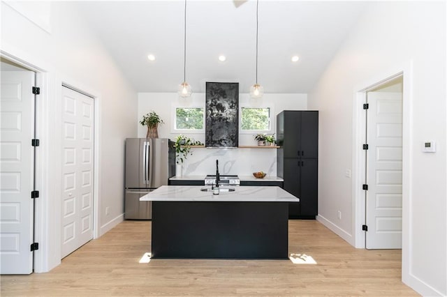 kitchen featuring decorative light fixtures, freestanding refrigerator, an island with sink, light wood finished floors, and light stone countertops