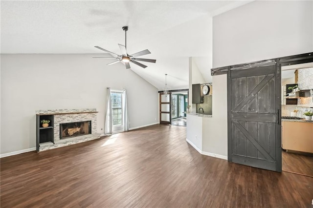 unfurnished living room with a fireplace with raised hearth, a ceiling fan, dark wood finished floors, a barn door, and baseboards