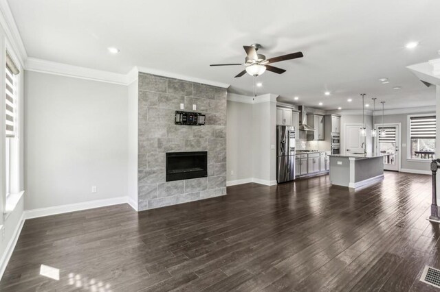 unfurnished living room featuring crown molding, dark wood-type flooring, ceiling fan, a healthy amount of sunlight, and a tiled fireplace