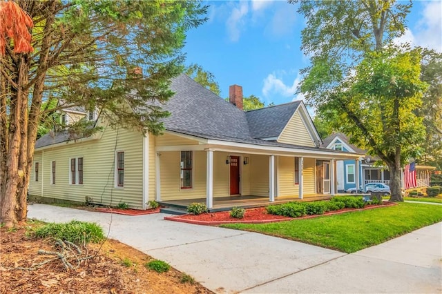 view of front facade featuring a chimney, a shingled roof, covered porch, a front yard, and driveway