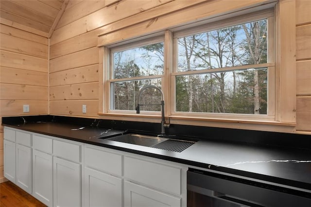 kitchen with dark countertops, wooden walls, a sink, white cabinets, and stainless steel dishwasher