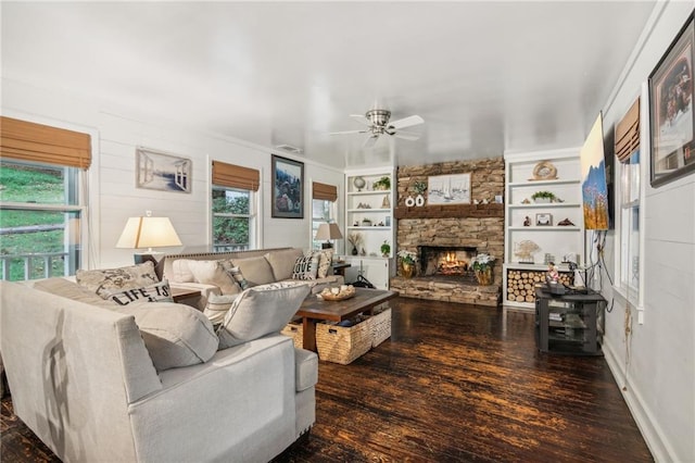 living room with ceiling fan, a stone fireplace, built in shelves, baseboards, and dark wood finished floors