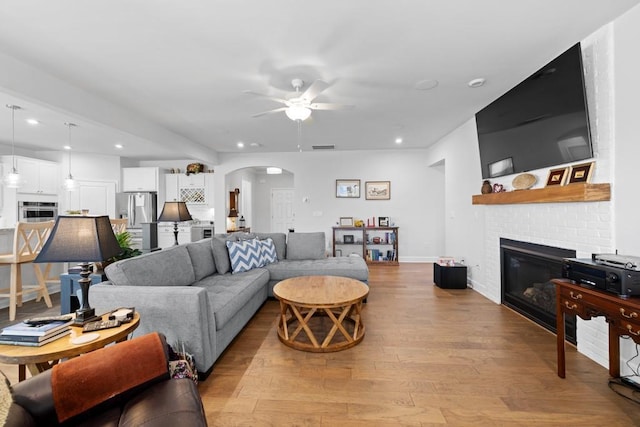 living room with light wood finished floors, recessed lighting, a fireplace, arched walkways, and ceiling fan