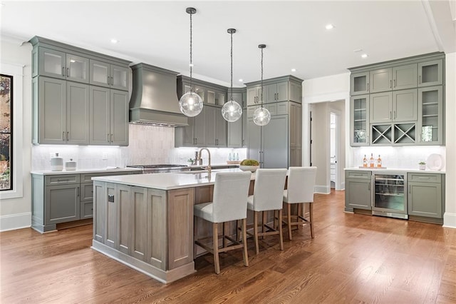 kitchen featuring a center island with sink, wine cooler, wood finished floors, custom exhaust hood, and light countertops