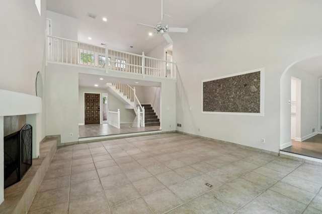 unfurnished living room featuring arched walkways, a ceiling fan, a fireplace with raised hearth, high vaulted ceiling, and stairs