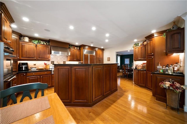 kitchen with backsplash, stainless steel double oven, a center island, wall chimney range hood, and light wood-type flooring