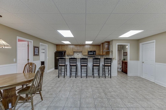 kitchen with light tile patterned floors, brown cabinetry, wainscoting, a kitchen breakfast bar, and black appliances