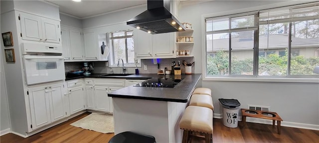 kitchen with dark countertops, a peninsula, a sink, white oven, and wall chimney range hood