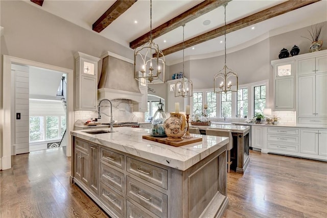 kitchen featuring hanging light fixtures, premium range hood, white cabinets, light stone countertops, and an island with sink