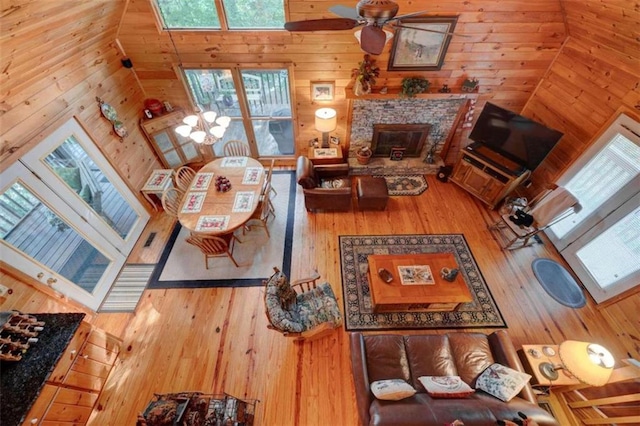 living room with ceiling fan, wooden walls, a stone fireplace, high vaulted ceiling, and wood-type flooring