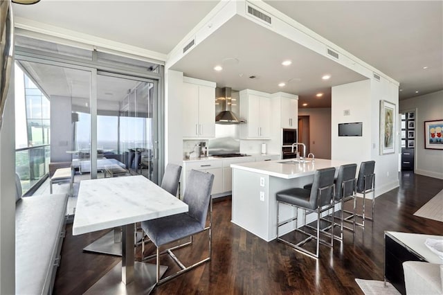 kitchen with white cabinetry, wall chimney range hood, gas stovetop, a kitchen breakfast bar, and an island with sink