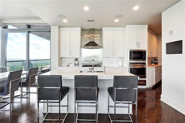 kitchen featuring stainless steel oven, a center island with sink, wall chimney exhaust hood, built in microwave, and white cabinetry